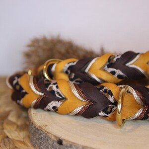May include: A braided collar featuring alternating strands of mustard yellow leather, dark brown leather, and a black and white leopard print fabric. The collar includes two polished gold-tone metal D-rings and a matching leather end tab.
