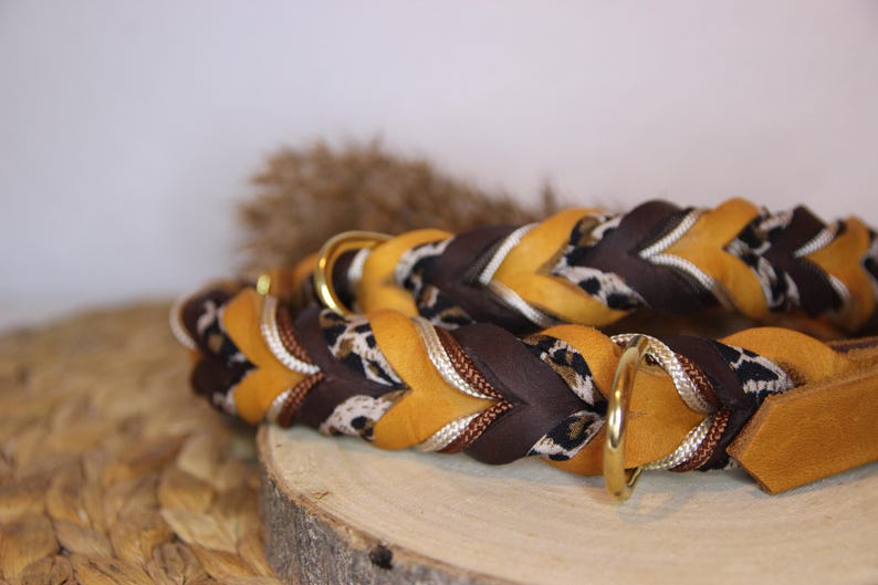 May include: A braided collar featuring alternating strands of mustard yellow leather, dark brown leather, and a black and white leopard print fabric. The collar includes two polished gold-tone metal D-rings and a matching leather end tab.