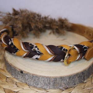 May include: A braided collar featuring alternating strands of mustard yellow, dark brown, and a white and black leopard print fabric. The collar includes a gold-toned metal D-ring and a matching gold-toned buckle closure with a tan leather strap end.