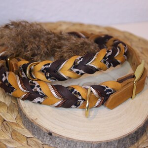 May include: A braided collar featuring alternating strands of mustard yellow, dark brown, and a black and white leopard print pattern. The collar includes gold-toned metal hardware, including a D-ring and a buckle, with a smooth leather end piece.