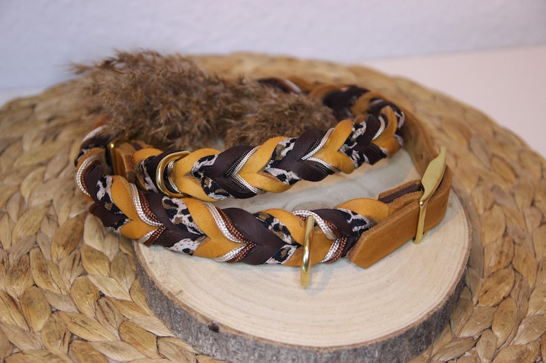 May include: A braided collar featuring alternating strands of mustard yellow, dark brown, and a black and white leopard print pattern. The collar includes gold-toned metal hardware, including a D-ring and a buckle, with a smooth leather end piece.