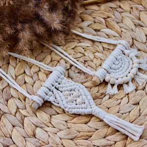 Puede incluir: Dos tapices de macramé de pared con listones de madera y cuentas blancas. Los tapices están hechos con cordón blanco y tienen detalles de flecos. Los tapices se muestran sobre una superficie de paja tejida.