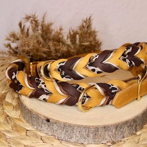 May include: A braided leather collar featuring alternating strands of mustard yellow, dark brown, and a black and white animal print pattern. The collar includes gold-toned metal hardware, including a D-ring and a buckle, with a smooth leather end.