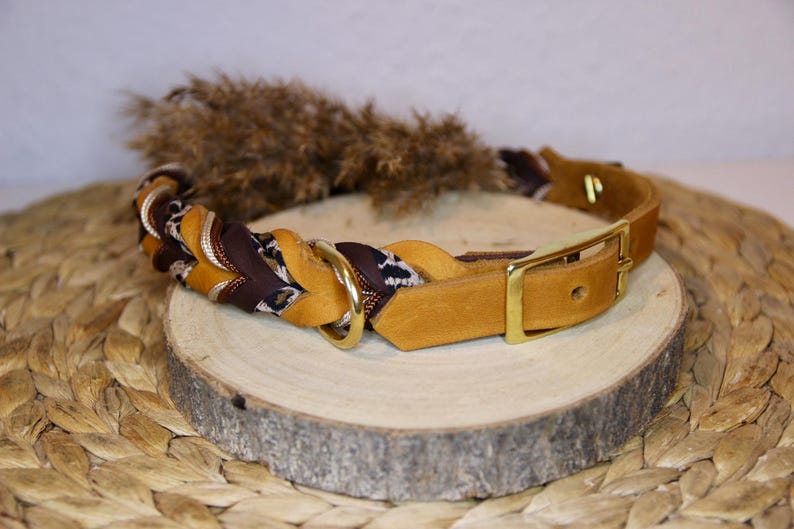 May include: A braided leather collar featuring a mix of mustard yellow, dark brown, and leopard print leather strands. The collar includes a gold-toned metal buckle, a matching D-ring for attachment, and a smooth leather strap end with a single hole.