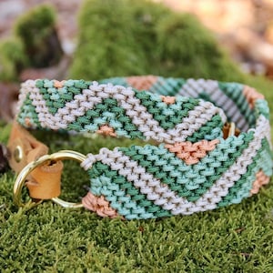 May include: A macrame dog collar with a geometric pattern in shades of green, peach, and cream. The collar features a brass ring and a leather strap. The collar is resting on a bed of green moss.