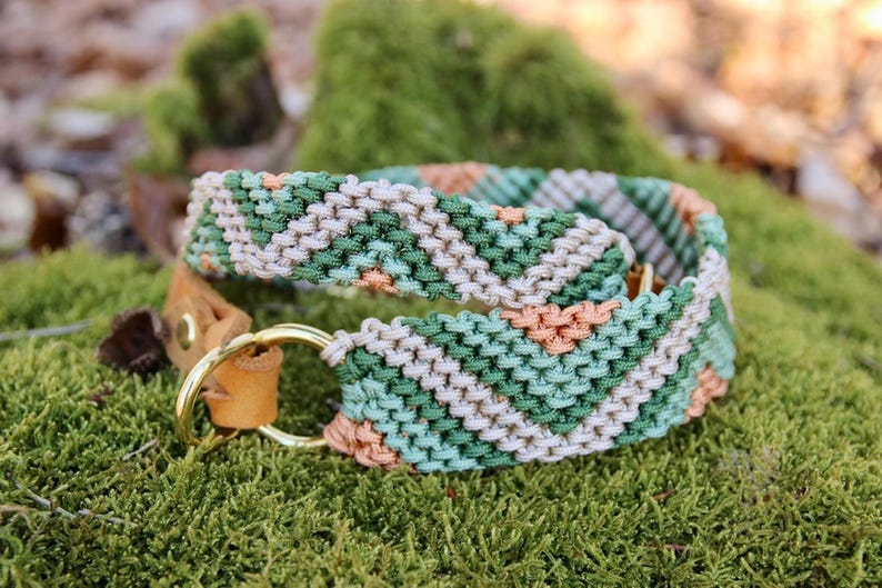 May include: A macrame dog collar with a geometric pattern in shades of green, peach, and cream. The collar features a brass ring and a leather strap. The collar is resting on a bed of green moss.