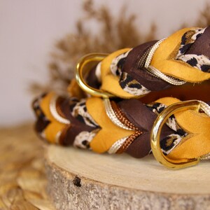 May include: A braided collar featuring mustard yellow and dark brown leather strips interwoven with a leopard print fabric and metallic gold cord. The collar includes a polished gold-tone metal D-ring for attachment, showcasing a multi-textured design.