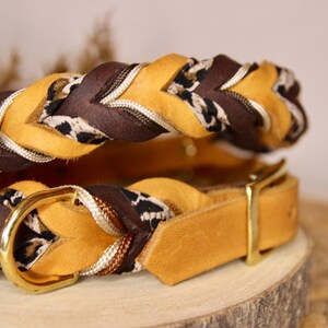 May include: A braided collar featuring mustard yellow and dark brown leather strips interwoven with a white and black leopard print fabric cord. The collar includes a polished gold-tone metal D-ring and a matching buckle fastener.