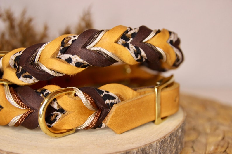 May include: A braided collar featuring mustard yellow and dark brown leather strips interwoven with a white and black leopard print fabric cord. The collar includes a polished gold-tone metal D-ring and a matching buckle fastener.