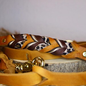 May include: A tan leather leash featuring a braided section composed of solid brown leather, tan leather, and a leopard print fabric strip. The leash includes gold-toned metal hardware, including a swivel snap hook and rivets.