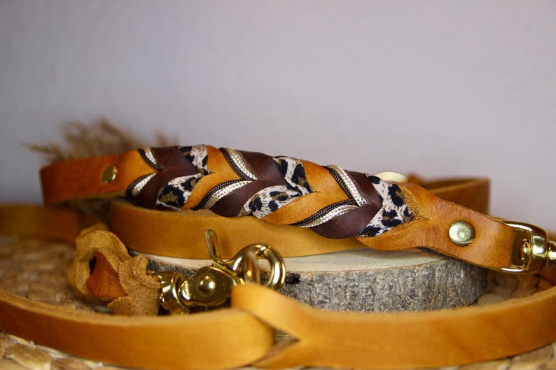 May include: A tan leather leash featuring a braided section composed of solid brown leather, tan leather, and a leopard print fabric strip. The leash includes gold-toned metal hardware, including a swivel snap hook and rivets.