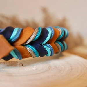 May include: A brown leather dog collar with a braided design featuring blue, grey, and tan cords. The collar has a silver metal D-ring.