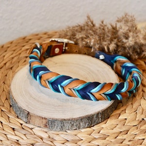 May include: A braided dog collar with brown leather and blue and teal ribbon. The collar is on a wooden round surface.