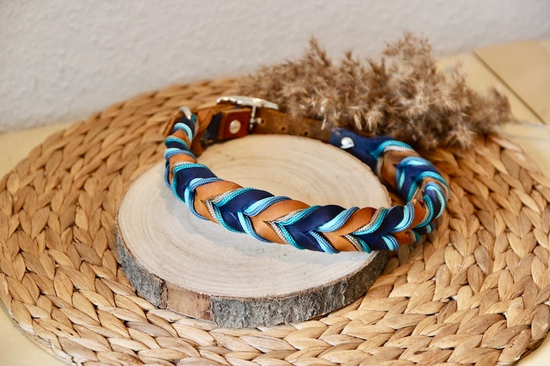May include: A braided dog collar with brown leather and blue and teal ribbon. The collar is on a wooden round surface.