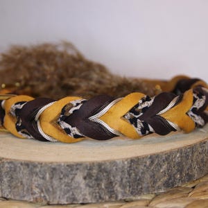 May include: A braided pet collar featuring alternating strands of mustard yellow, dark brown, and a black and white leopard print pattern. The collar includes a gold-toned metal D-ring for attachment and is constructed with a thick, woven design.