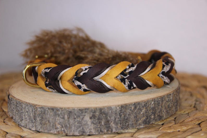 May include: A braided pet collar featuring alternating strands of mustard yellow, dark brown, and a black and white leopard print pattern. The collar includes a gold-toned metal D-ring for attachment and is constructed with a thick, woven design.