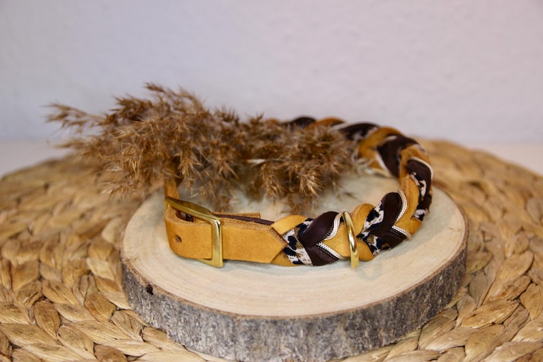 May include: A braided collar featuring tan leather, dark brown fabric, and a white and black leopard print pattern. The collar includes a gold-toned metal buckle and a D-ring attachment, with a smooth leather strap end and a metal pin closure.