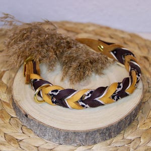 May include: A braided pet collar featuring alternating strands of mustard yellow, dark brown, and a black and white patterned fabric. The collar includes a gold-toned metal D-ring and a matching gold-toned metal stud fastener for secure closure.