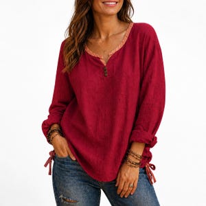 May include: A long-sleeved red blouse with a V-neckline and button details. The blouse has a contrasting peach-colored trim around the neckline and cuffs. The sleeves are rolled up, and the sides are tied with a ribbon. The model is wearing blue jeans.