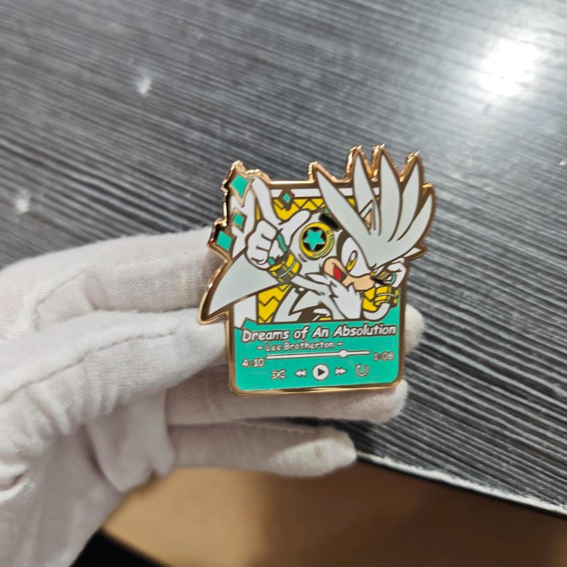 May include: A gold-colored metal pin featuring a cartoon character with white fur and spiky hair. The pin has a rectangular design with the text "Dreams of An Absolution" and music player controls. The character is set against a yellow and teal background.