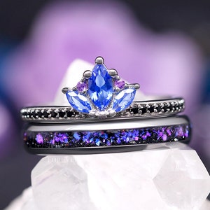 May include: A black and silver ring set with a blue gemstone surrounded by smaller purple gemstones. The ring is on a white crystal surface.