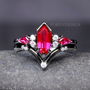 May include: Black ring set with a large, red, octagon-shaped gemstone and smaller red gemstones on either side. The ring is set with white gemstones and has a V-shaped band. The text "FOREVERINGS" is visible in the image.