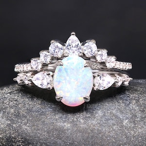May include: A silver ring set with a large oval opal surrounded by smaller clear stones. The ring is on a gray rock.