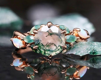 May include: Rose gold ring featuring a large, round moss agate center stone with green inclusions. The ring is accented with smaller green stones and opals. The ring is set against a dark, reflective surface.