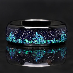 May include: A black ring with a dark blue interior speckled with glitter and a band of teal-colored stones. The ring is highly reflective, with a mirrored image of itself on the surface below.