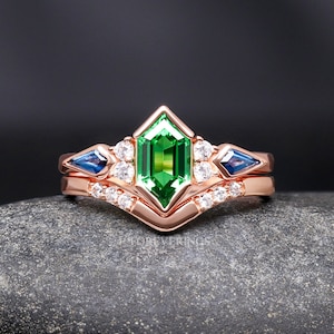 Könnte beinhalten: Ein Verlobungsring aus Roségold mit einem grünen Smaragdschliff-Edelstein, der von kleineren weißen Edelsteinen umgeben ist. Die Band hat ein Chevron-Design und zwei blaue Saphir-Akzentsteine.