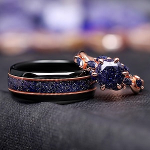 May include: A pair of rings, one a black band with a blue speckled inlay and copper-colored trim, the other a rose gold ring with a large blue stone and smaller blue accent stones. The rings are set against a dark background.