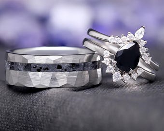 Op de afbeelding: Twee trouwringen. Eén is een zilveren band met een getextureerd, geometrisch ontwerp en een donkergrijze inleg. De andere is een zilveren ring met een zwarte, peervormige steen, omringd door kleinere witte stenen. De ringen liggen op een donkergrijze ondergrond.