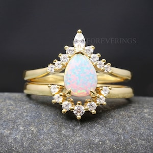 May include: A gold ring set with a pear-shaped opal and a halo of smaller clear stones. The ring is on a gray background.