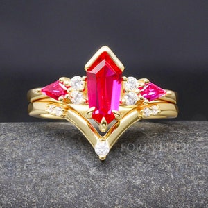 Peut inclure: Une bague en or sertie d'une grande pierre précieuse rectangulaire rose vif entourée de petites pierres précieuses claires. La bague est posée sur une surface gris foncé.