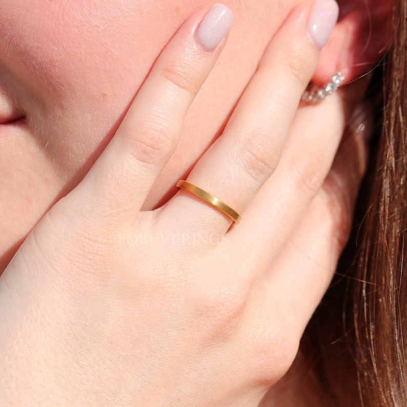 May include: A gold-colored ring with a flat, rectangular band. The ring is on a finger, with the text "FOREVERRINGS" visible in the background. The ring appears to be made of metal and has a simple, elegant design.