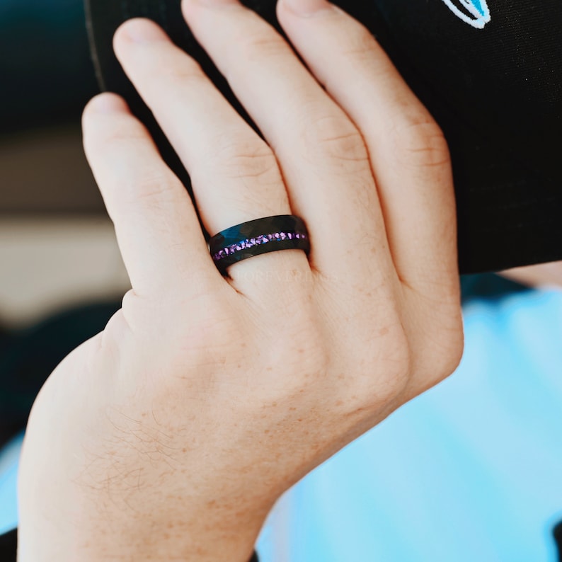 May include: A black ring with a row of purple gemstones. The ring is worn on the finger of a person's hand.