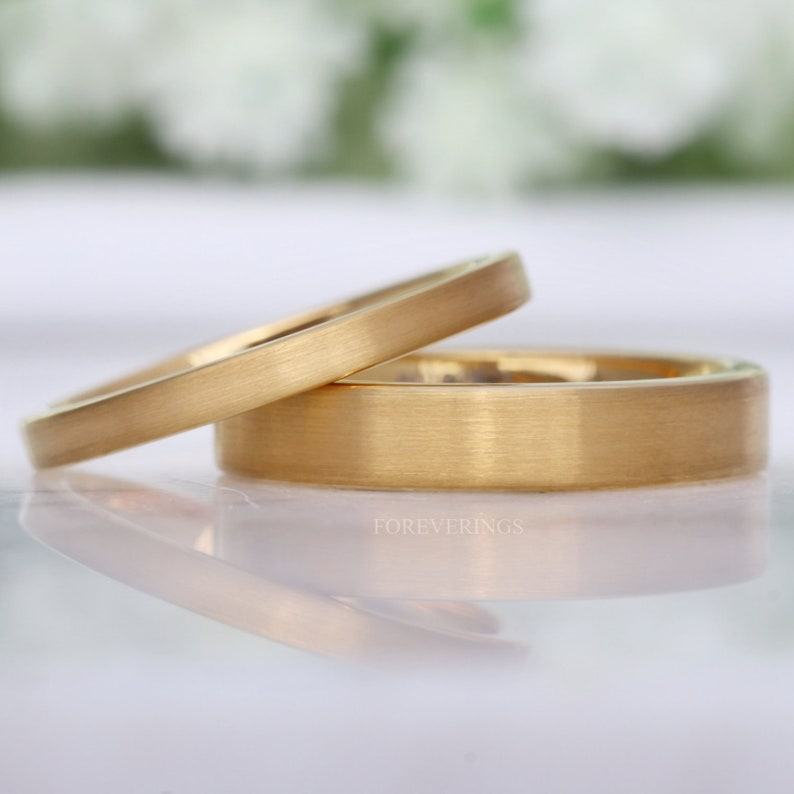 May include: Two gold wedding bands with a brushed finish. One ring is slightly angled, resting on top of the other. The rings have a flat profile and a matte appearance. The word "FOREVERRINGS" is visible.