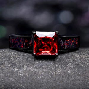 May include: A black ring with a large, square, red gemstone as the centerpiece. The band features a dark, speckled design with hints of red and purple. The ring is set against a dark, textured background.