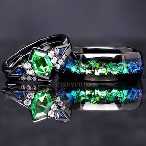 May include: A black wedding band set with a green emerald gemstone and blue sapphire accents. The band is set with small clear diamonds. The other band is black with green and blue glitter.