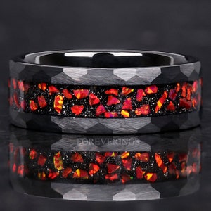 May include: A black ring with a hammered texture and a band of red and orange gemstones set in a black speckled background. The ring has a polished, reflective surface.
