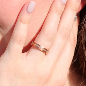 May include: A rose gold wedding band with a hammered finish and a thin white gold inlay. The band is worn on a finger.