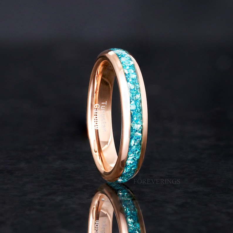 May include: Rose gold ring with a turquoise gemstone band. The ring is engraved with the words "Tungsten Carbide".