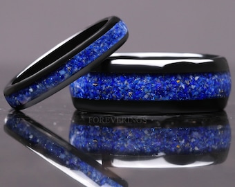 Può includere: Due anelli neri con un'intarsio blu vibrante. L'intarsio è fatto di pietra frantumata con scaglie d'oro. Gli anelli sono esposti su una superficie riflettente. Il testo "FOREVERRINGS" è visibile.