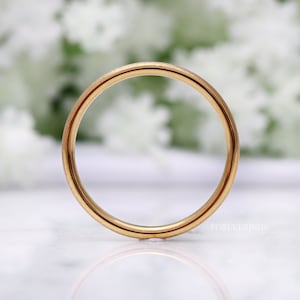 May include: A simple gold wedding band with a smooth, polished finish. The band is shown on a white surface with a blurred background.
