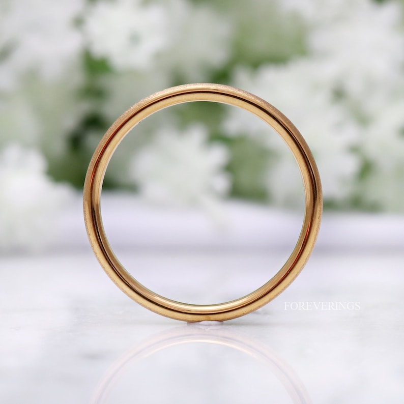 May include: A simple gold wedding band with a smooth, polished finish. The band is shown on a white surface with a blurred background.