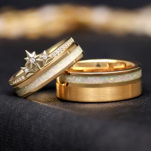 Könnte beinhalten: Zwei goldene Eheringe. Ein Ring hat ein Sterndesign mit Diamanten und einen weißen Opal-Inlay. Der andere Ring ist ein schlichtes goldenes Band mit einem weißen Opal-Inlay. Die Ringe liegen auf einer dunklen Oberfläche.