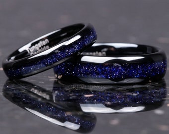 Può includere: Due anelli neri in carburo di tungsteno con un'incisione di glitter blu. Gli anelli sono impilati l'uno sull'altro e si riflettono su una superficie lucida.