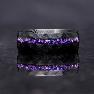 May include: A black carbon fiber ring with a band of purple and blue crushed glass inlay. The ring is reflecting on a dark surface.