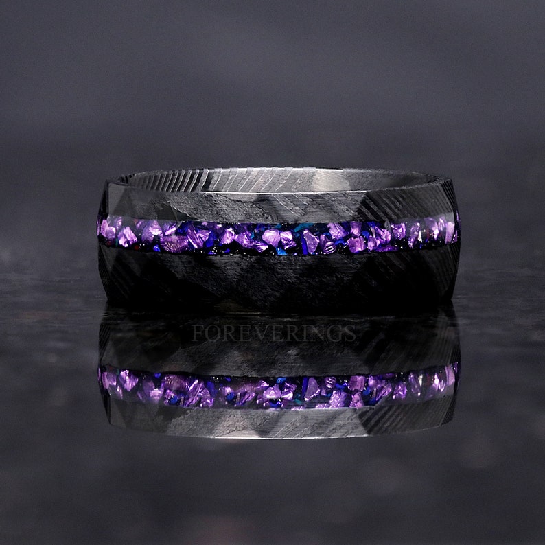 May include: A black carbon fiber ring with a band of purple and blue crushed glass inlay. The ring is reflecting on a dark surface.
