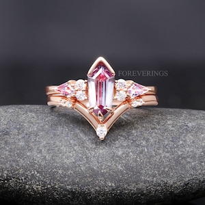 May include: A rose gold engagement ring set with a large, rectangular, purple and pink gemstone surrounded by smaller, round, white gemstones. The ring is set on a gray, textured surface. The text "FOREVERINGS" is visible in the upper right corner of the image.